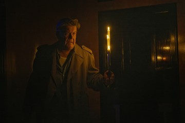 Pimeässä huoneessa takkiin pukeutunut mies pitää sytytettyä kynttilää, hänen kasvonsa ovat kireät ja kynttilänvalo valaisee hänen seisoessaan suljetun oven lähellä.