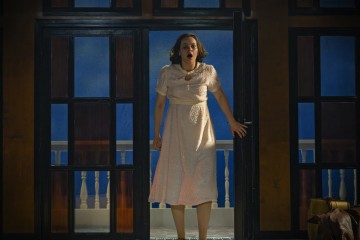 Vaaleanpunaiseen mekkoon pukeutunut nainen seisoo oviaukossa, pitelee ovea toisella kädellään ja näyttää järkyttyneeltä. Taustalla on siniset seinät ja valkoinen kaide, ja hänen vieressään lattialla on matkalaukku.