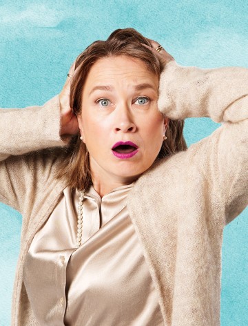 Vaaleanruskeat hiukset ja kirkkaan vaaleanpunainen huulipuna omaava nainen katsoo yllättyneenä, silmät suurina ja suu auki, ja pitää käsiään päänsä päällä. Hänellä on yllään beige pusero ja satiinipusero, ja hän seisoo sinistä taustaa vasten.
