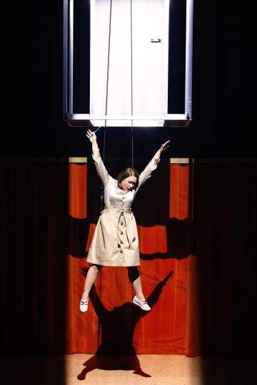 Beigeen trenssitakkiin pukeutunut nainen roikkuu käsistään kiinni suorakulmaisesta kehyksestä, jalat roikkuvat hänen yläpuolellaan, punaisten verhojen ja dramaattisen valaistuksen edessä, joka heittää suuren varjon hänen takanaan olevalle seinälle.