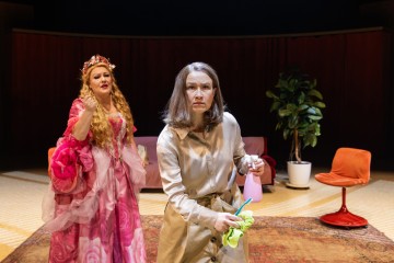 Kaksi naista esiintyy lavalla. Toisella on vaaleanpunainen puku ja kruunu, ja hän näyttää dramaattiselta; toinen on pukeutunut beigeen ja näyttää keskittyneeltä pitäessään kädessään siivoustarvikkeita. Lavastukseen kuuluu kasvi ja oransseja tuoleja taustalla.