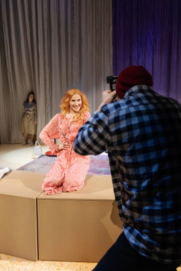 Vaaleanpunaiseen mekkoon pukeutunut nainen poseeraa ja hymyilee valokuvaajalle, jolla on ruudullinen paita ja punainen pipo. Toinen nainen seisoo taustalla lähellä verhoja ja rekvisiittaa.