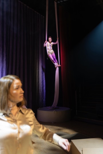 Hohtavaan pukuun pukeutunut ilmataiteilija roikkuu lavalla valkoisista silkkeistä, kun taas beigeen puseroon pukeutunut nainen seisoo epäselvässä etualalla dramaattisessa valaistuksessa.