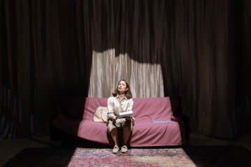 Nainen istuu yksin violetilla sohvalla auringonvalossa, paperit sylissään ja beige käsilaukku vieressä. Hämärässä huoneessa näkyvät raskaat verhot ja kuviollinen matto.