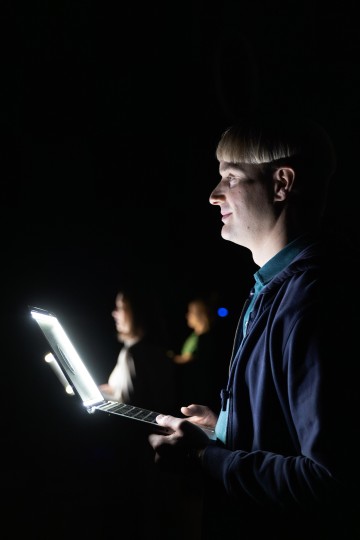 Lyhyet vaaleat hiukset omaava henkilö seisoo pimeässä huoneessa, jota valaisee avoimen kannettavan tietokoneen valo. Taustalla näkyy kaksi epäselvää hahmoa, jotka ovat osittain valaistuja.