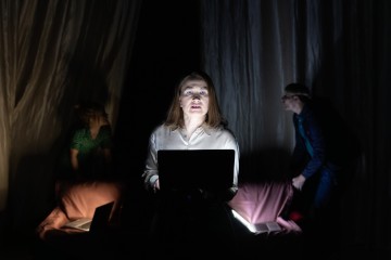 Nainen seisoo hämärässä valossa kannettava tietokone kädessään ja katsoo eteenpäin. Taustalla on kaksi ihmistä, jotka ovat osittain peitossa ja joilla on myös kannettavat tietokoneet. Kohtaus on dramaattinen varjojen ja verhojen avulla.