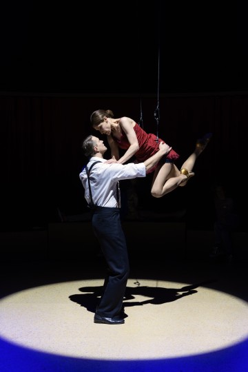 Viralliseen asuun pukeutunut mies pitelee punamekkoon pukeutunutta naista, joka heiluu trapetsilla valonheittimen alla, ja tumma tausta viittaa teatteri- tai sirkusesitykseen.