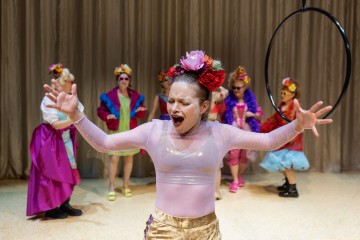 Henkilö, jolla on kiiltävä vaaleanpunainen toppi ja kultaiset shortsit, laulaa intohimoisesti kädet levällään, ja hänellä on värikäs kukkainen päähine. Taustalla tanssii kultaisen verhon edessä viisi muuta, joilla on samanlainen juhlava asu ja asusteet.