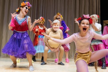 Joukko värikkäisiin, omituisiin pukuihin ja peruukkeihin pukeutuneita aikuisia esiintyy lavalla energisesti, iloisin ilmein ja liioitelluissa asennoissa beigen verhon edessä.