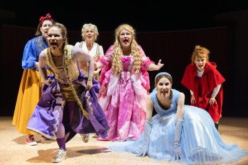 Kuusi värikkäisiin satupukuihin pukeutunutta naista esiintyy lavalla ja ilmaisee voimakkaita tunteita. Heillä on päällään palloilupuvun, viitan tai hansikkaiden kaltaisia asuja, ja he näyttävät olevan keskellä toimintaa, mahdollisesti laulavat tai huutavat dramaattisessa kohtauksessa.