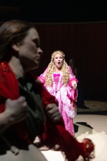 Vaaleanpunaiseen, koristeelliseen mekkoon pukeutunut nainen, jolla on pitkät vaaleat kiharat, seisoo näyttämön keskellä suu auki järkyttyneenä. Etualalla on epäselvästi toinen nainen punaisessa huivissa kävelemässä poispäin. Tausta on hämärästi valaistu.