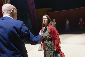 Vihreään mekkoon ja punaiseen huiviin pukeutunut nainen seisoo lavalla kaljua pukumiestä vastapäätä ja näyttää järkyttyneeltä. Taustalla seisoo kolme mekkoon pukeutunutta naista dramaattisessa näyttämövalaistuksessa.