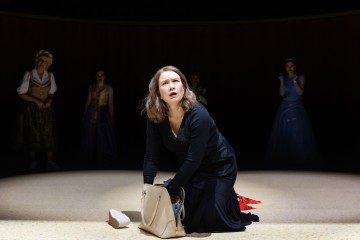 Mustapukuinen nainen polvistuu lavalla ja katsoo ylöspäin huolestuneena. Hänellä on kädessään avonainen laukku, jonka vieressä on valkoinen kenkä. Taustalla seisoo neljä epäselvää hahmoa, jotka katselevat häntä.