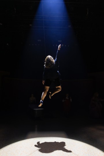 Tummiin vaatteisiin pukeutunut henkilö leijuu ilmassa dramaattisen valonheittimen alla, kurottaen yhdellä kädellään ylöspäin ja heittäen selkeän varjon lattialle pimeässä teatterissa.