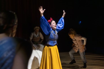 Lumikki-pukuun pukeutunut nainen seisoo näyttämön keskellä kädet kohotettuna ja näyttää ahdistuneelta, kun taas kaksi muuta pukuun pukeutunutta näyttelijää reagoi dramaattisesti hänen takanaan teatterivalaistuksessa.