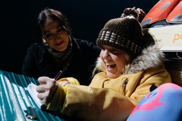 Hymyilevä tyttö keltaisessa takissa ja ruskeassa neulomyssyssä kirjoittaa muistikirjaan, kun aikuinen nainen katselee lämpimästi. He ovat ulkona yöllä matkalaukkujen ja laukkujen vieressä.