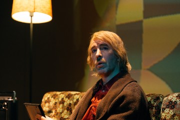 Mies, jolla on olkapäiden pituinen vaalea tukka ja viikset, istuu kuviollisella sohvalla ja pitelee kehystettyä valokuvaa. Hänellä on ruskea villapaita ja punainen paita. Hänen vieressään seisoo sytytetty lamppu, joka heittää lämpimän valon hämärään huoneeseen.