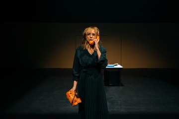 Tummapukuinen nainen seisoo hämärästi valaistulla näyttämöllä, pitää oranssia johdollista puhelinta korvallaan ja katsoo eteenpäin. Toisessa kädessään hän pitää puhelimen jalustaa, ja hänen takanaan on pieni tumma pöytä ja kirjoja.