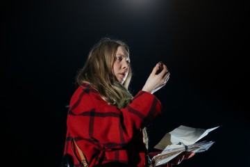 Punaiseen ruututakkiin pukeutunut nainen seisoo tummaa taustaa vasten, kädessään paperipino ja katselee tarkasti kädessään olevaa pientä esinettä, jota valonheitin valaisee.