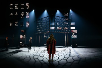 Pitkään punaiseen takkiin pukeutunut henkilö seisoo kuusikulmaista metallirakennetta vasten, ja dramaattinen valaistus heittää lattialle geometrisia varjoja. Kaksi muuta ihmistä seisoo erillään hämärästi valaistulla näyttämöllä, jonka taustalla on digitaalisia käsikirjoitusprojektioita.