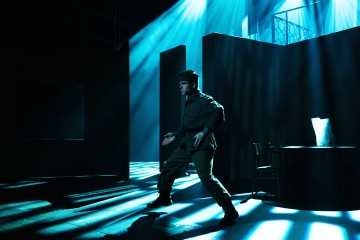 Sotilaspukuinen mies seisoo dramaattisessa poseerauksessa hämärästi valaistulla näyttämöllä, jonne virtaa sinisiä valonsäteitä. Taustalla näkyy varjoisia lavasteita ja pöytä, jossa on valkoinen kassi.