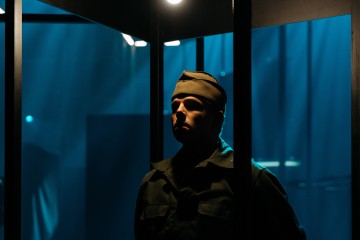Sotilaspukuinen ja lippalakki päässä oleva henkilö seisoo hämärässä, sinisellä valaistussa huoneessa, jota osittain kehystävät tummat metallirimat ja joka on valaistu ylhäältä.