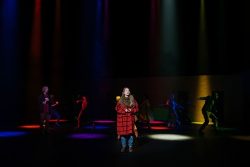 Nuori nainen punaisessa ruudullisessa takissa seisoo näyttämön keskellä kirkkaiden, värikkäiden valonheittimien alla, kun taas muut esiintyjät liikkuvat taustalla, osittain punaisen, keltaisen, sinisen ja vihreän valon säteiden valaisemina.