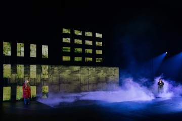 Kaksi ihmistä seisoo erillään sumuisella näyttämöllä, toinen punatakkinen vasemmalla ja toinen tummiin vaatteisiin pukeutunut oikealla. Heidän takanaan on valaistuja suorakaiteen muotoisia paneeleita, joissa on vihertäviä kuvioita, ja sivusta loistaa valonheitin.