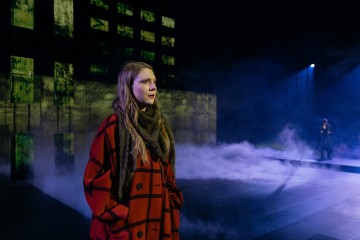 Punaruutuinen nainen seisoo sumun ympäröimällä hämärästi valaistulla näyttämöllä mietteliään näköisenä. Taustalla toinen henkilö seisoo vihreistä ja mustista paneeleista koostuvan seinän vieressä.