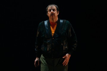 Mies, jolla on silmälasit, tumma kuviollinen takki ja oranssi paita, seisoo dramaattisessa valaistuksessa tummaa taustaa vasten ja katsoo hieman ylöspäin vakavalla ilmeellä.