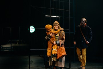 Talvitakkiin ja neulomyssyyn pukeutunut nainen halaa lasta, jolla on oranssi lumipuku. Lähistöllä seisoo mies, jolla on tumma takki ja keltaiset housut. He ovat hämärästi valaistulla näyttämöllä, jossa on minimaalinen lavastus ja taustalla auto.