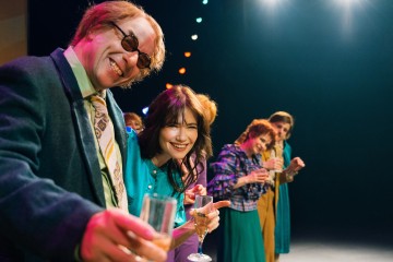 Ryhmä värikkäisiin retro-vaatteisiin pukeutuneita ihmisiä seisoo lavalla rivissä hymyillen ja juomia kädessään, ja heidän yläpuolellaan loistavat kirkkaat lavavalot.