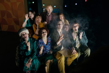 Yhdeksän ihmisen ryhmä, enimmäkseen retrovaatteisiin pukeutuneena, istuu tiiviisti yhdessä penkillä, pitelee savukkeita kädessään ja katsoo ylöspäin ilmeikkäillä kasvoilla, kun heidän yläpuolellaan roikkuu värikkäitä valonjohtoja. Savu leijailee ilmassa.