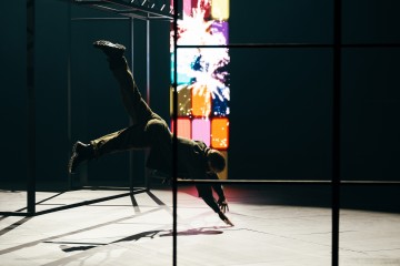 Henkilö tekee dynaamisen akrobaattisen liikkeen tasapainoilemalla vaakatasossa yhdellä kädellä metallirakenteen päällä. Taustalla näkyy elävä, monivärinen digitaalinen näyttö, jossa on abstrakteja kuvioita ja ilotulituksia.