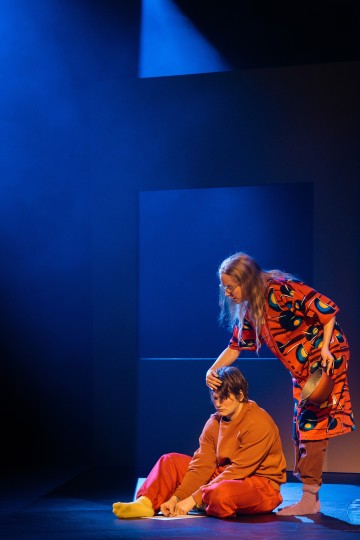 Kaksi näyttelijää esiintyy hämärästi valaistulla näyttämöllä. Toinen istuu lattialla oransseissa vaatteissa ja katsoo surullisena alaspäin. Toinen seisoo värikkäässä kaapussaan ja laskee kätensä varovasti istuvan näyttelijän pään päälle, ja sininen valo valaisee kohtausta.