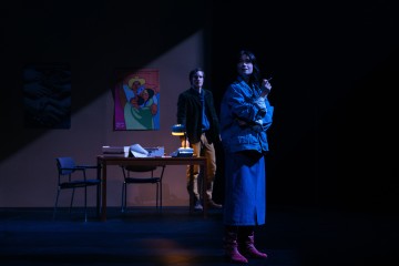 Farkkutakkinen ja vaaleanpunaisiin saappaisiin pukeutunut nainen seisoo valonheittimen alla savuke kädessään. Hänen takanaan mies seisoo pöydän ääressä, jossa on lamppu, papereita ja tuoleja, ja seinällä on värikkäitä julisteita. Ympäristö on hämärästi valaistu.