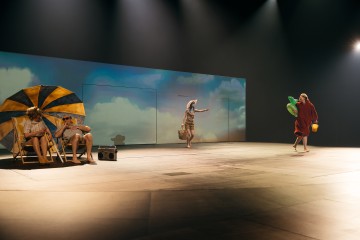 Neljä ihmistä näyttelee lavalla rantakohtausta; kaksi istuu keltaisen ja sinisen sateenvarjon alla radion kanssa, yksi seisoo laukku ja hattu kädessään, ja toinen kävelee punaisessa pyyhkeessä puhallettavien lelujen ja ämpärin kanssa.