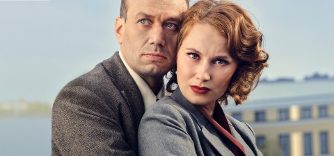 Vakava mies ja nainen, joilla molemmilla on harmaa puku, seisovat lähekkäin ulkona. Naisella on lyhyet, aaltoilevat punaiset hiukset ja rohkea meikki. Molemmat katsovat keskittyneesti kameraan, jonka taustalla on epäselvä rakennus.