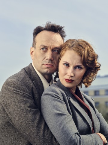 Vintage-tyylisiin pukuihin pukeutuneet mies ja nainen seisovat lähekkäin ulkona, ja molemmat katsovat vakavasti kameraan. Naisella on kiharat punaruskeat hiukset ja miehellä lyhyet tummat hiukset. Taustalla näkyy sumea rakennus ja taivas.