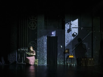 Pitkään vaaleanpunaiseen mekkoon pukeutunut nainen istuu pienellä penkillä lukemassa kirjaa hämärästi valaistulla näyttämöllä tiiliseinän, vintage-gramofonin ja matkalaukkujen läheisyydessä; dramaattiset varjot ja valonheittimet luovat tunnelmallisen tunnelman.