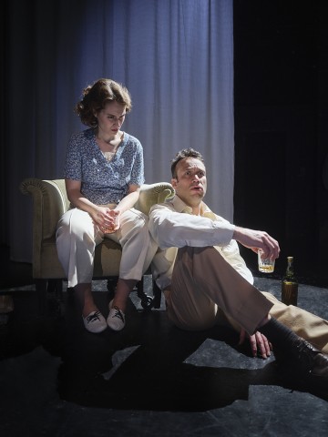 Nainen istuu pienellä sohvalla drinkki kädessään ja katsoo vakavana alaspäin. Mies istuu lattialla naisen vieressä nojaillen taaksepäin, hänellä on myös juoma kädessään ja pullo vieressä. Kohtaus on hämärästi valaistu ja dramaattinen.