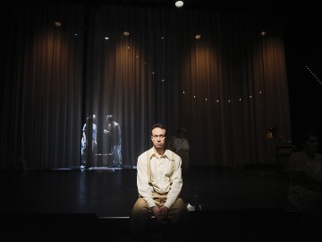 Vaaleisiin vaatteisiin pukeutunut henkilö istuu näyttämön keskellä, valonheittimen valaisemana, ja kaksi hahmoa näkyy heikosti verhon takana, ja taustalla on hämärät valonjohdot, jotka luovat dramaattisen teatterikohtauksen.