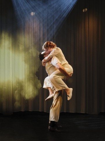 Mies nostaa hymyilevän naisen syliinsä hämärästi valaistulla näyttämöllä, jossa valonheittimet loistavat ja savu luo dramaattisen vaikutelman. Molemmat ovat pukeutuneet vintage-tyylisiin vaaleisiin vaatteisiin.