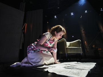 Nuori nainen, jolla on kukkakuvioinen esiliina, polvistuu näyttämön lattialla ja kirjoittaa keskittyneesti edessään levittäytyneille sanomalehden sivuille, taustalla vihreä nojatuoli ja dramaattinen näyttämövalaistus.