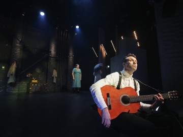 Valkoiseen paitaan ja sukkanauhoihin pukeutunut mies soittaa klassista kitaraa lavalla, jota valot valaisevat. Taustalla seisoo siniseen mekkoon pukeutunut nainen, jonka takana on tumma, teatterimainen lavaste.