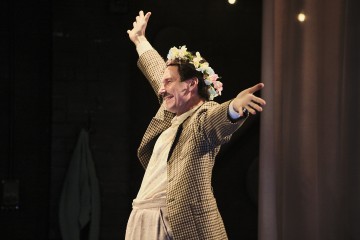 Henkilö, jolla on kukkakruunu ja ruudullinen takki, seisoo iloisesti kädet ojennettuina ja hymyilee lämpimässä näyttämövalaistuksessa.