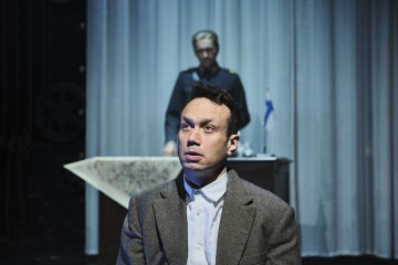 Etualalla istuu pukumies huolestunut ilme kasvoillaan, kun taas toinen sotilaspukuinen mies seisoo taustalla pöydän takana. Valkoinen verho ja pieni lippu ovat näkyvissä.