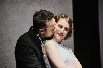 Tummapukuinen mies syleilee ja suutelee hymyilevää kiharatukkaista naista, jolla on hihaton valkoinen mekko. He seisovat lähekkäin harmaata taustaa vasten, ja nainen katsoo sivulle.