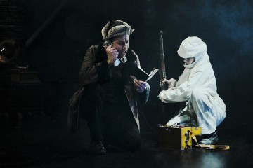 Henkilö, jolla on turkislakki ja -takki, puhuu vintage-puhelimeen ja lukee samalla muistikirjaa, kun toinen, valkoisiin suojavaatteisiin pukeutunut henkilö polvistuu lähistöllä ja käsittelee kivääriä ja keltaista salkkua pimeällä, savuisella näyttämöllä.