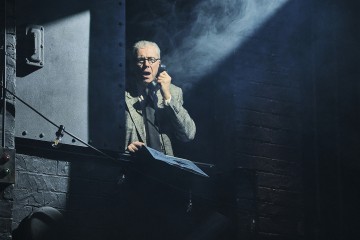 Pukuun ja silmälaseihin pukeutunut vanhempi mies seisoo dramaattisessa valaistuksessa metalliseinän vieressä, puhuu vanhaan puhelimeen ja pitää kädessään papereita, ja hänen ympärillään pyörteilee savua pimeässä teollisuusympäristössä.